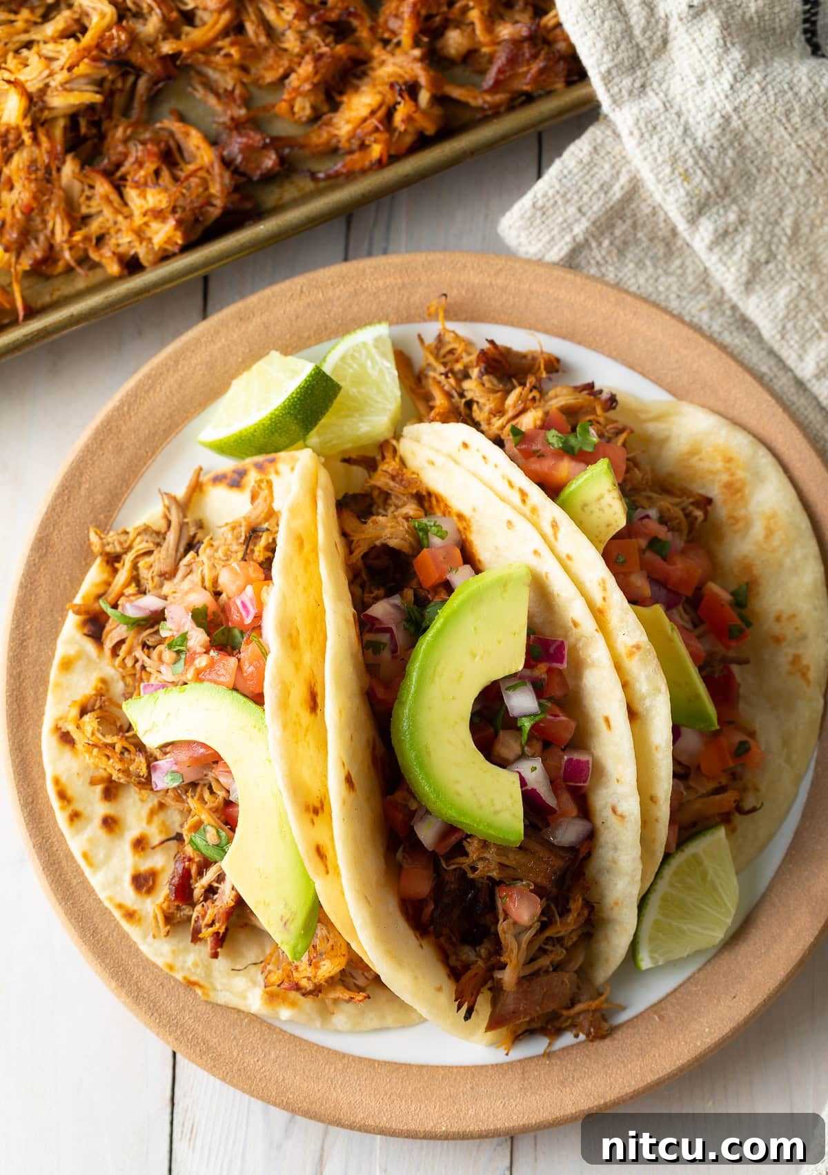 Overhead shot of three perfectly assembled carnitas tacos with avocado slices on a plate, ready to be enjoyed.
