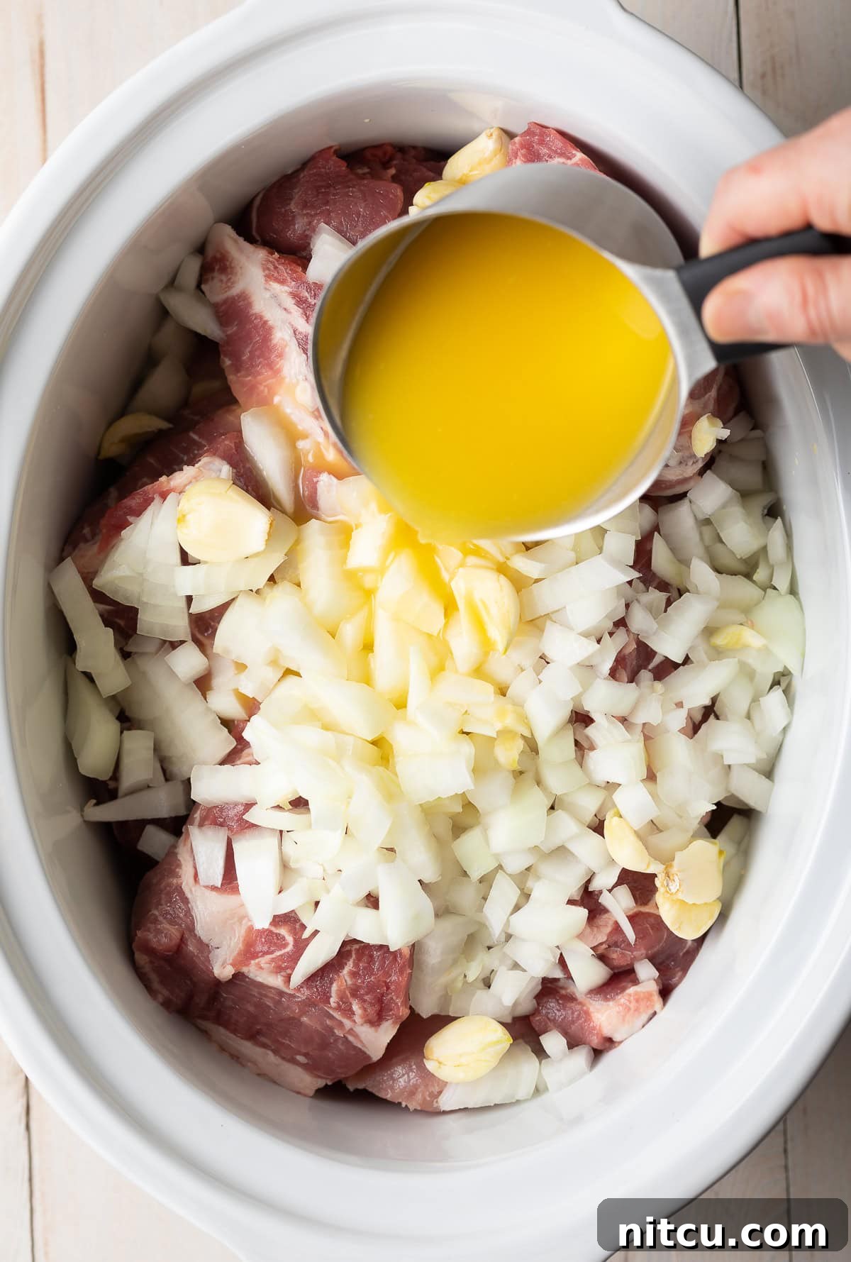 A hand pouring a cup of fresh orange juice into a slow cooker filled with pork, garlic, and onions.