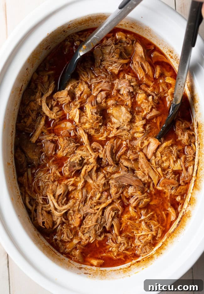 Tongs gently shredding the perfectly cooked carnitas inside the slow cooker, submerged in its flavorful juices.