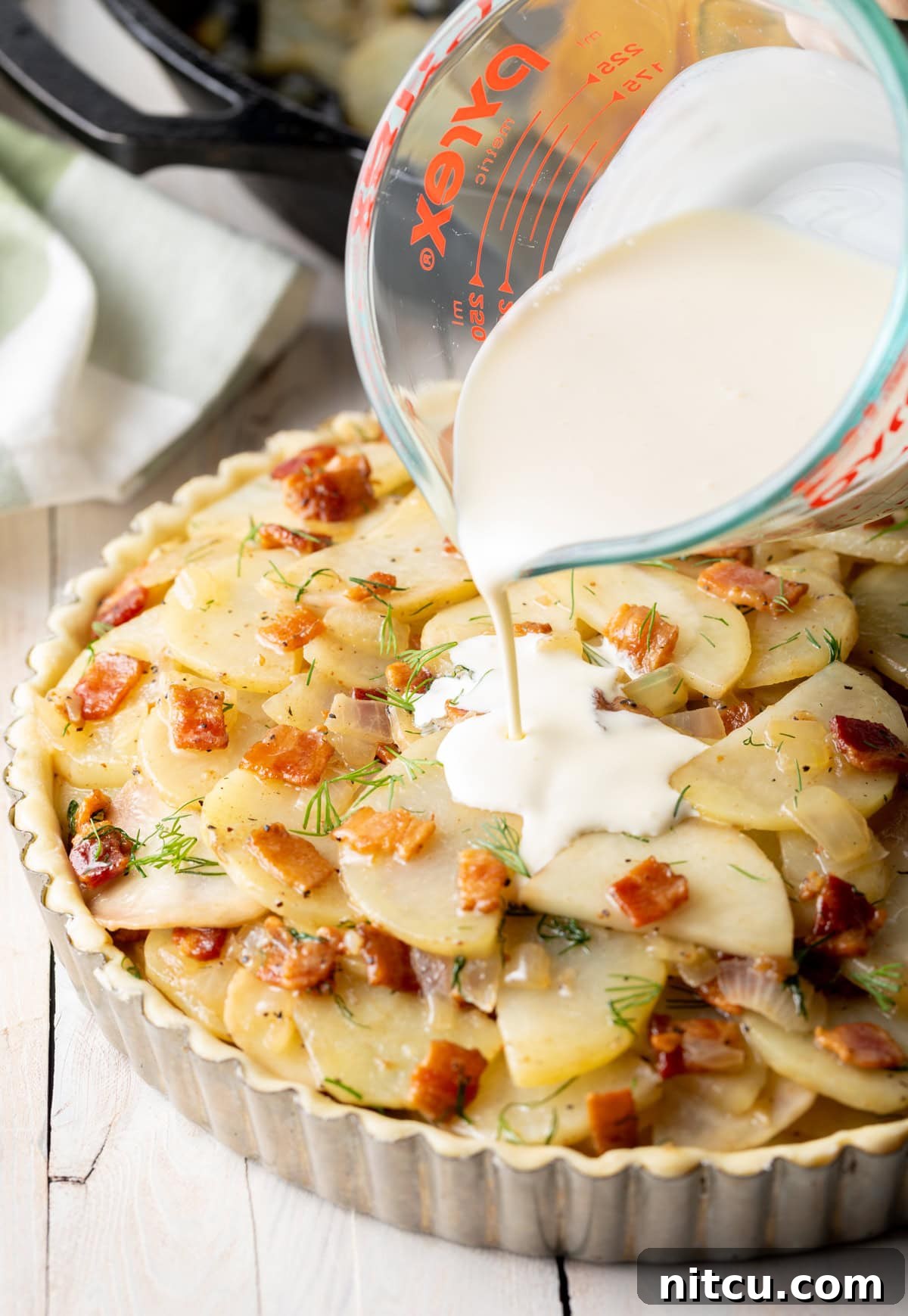A rich stream of heavy cream being poured over the layered potatoes and bacon mixture inside a tart tin, preparing for baking.