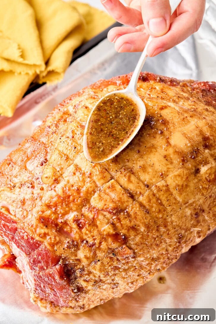 A close-up view of a ham on a baking sheet, with a generous layer of brown sugar glaze being brushed over its scored surface, preparing it for baking to achieve a glossy, caramelized finish.