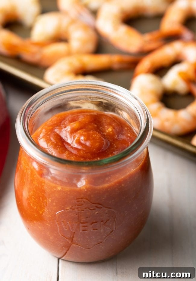 Small jar of homemade cocktail sauce recipe with succulent shrimp in the background, ready to be dipped.