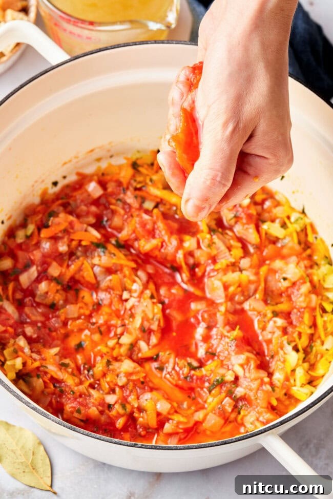 A hand squishing a whole peeled San Marzano tomato over a pot, breaking it into irregular pieces for the soup.