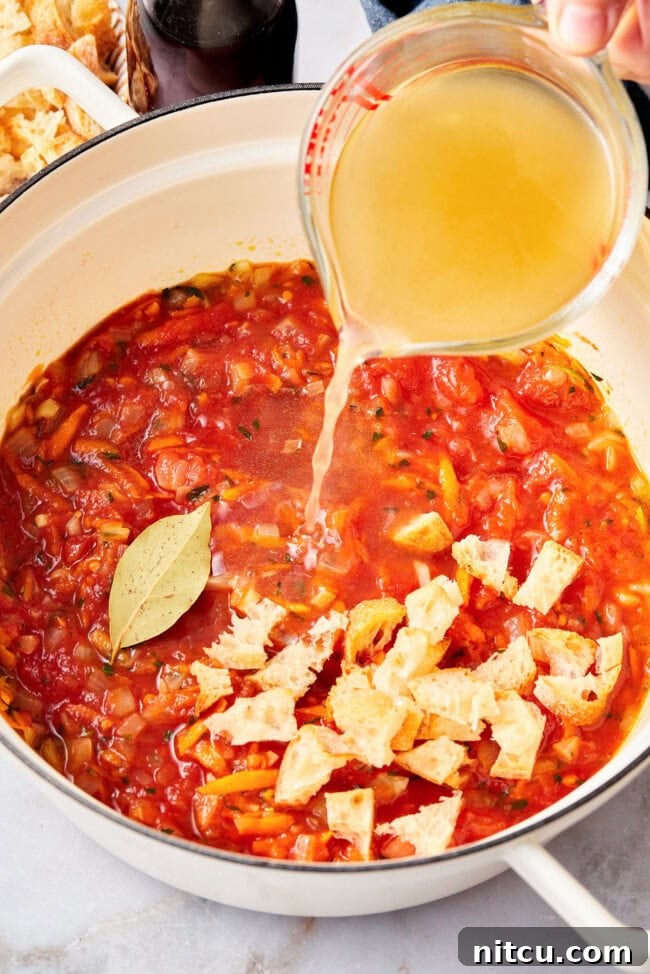 Vegetable broth being poured into a large stockpot filled with tomatoes, vegetables, and bread, for making tomato soup.