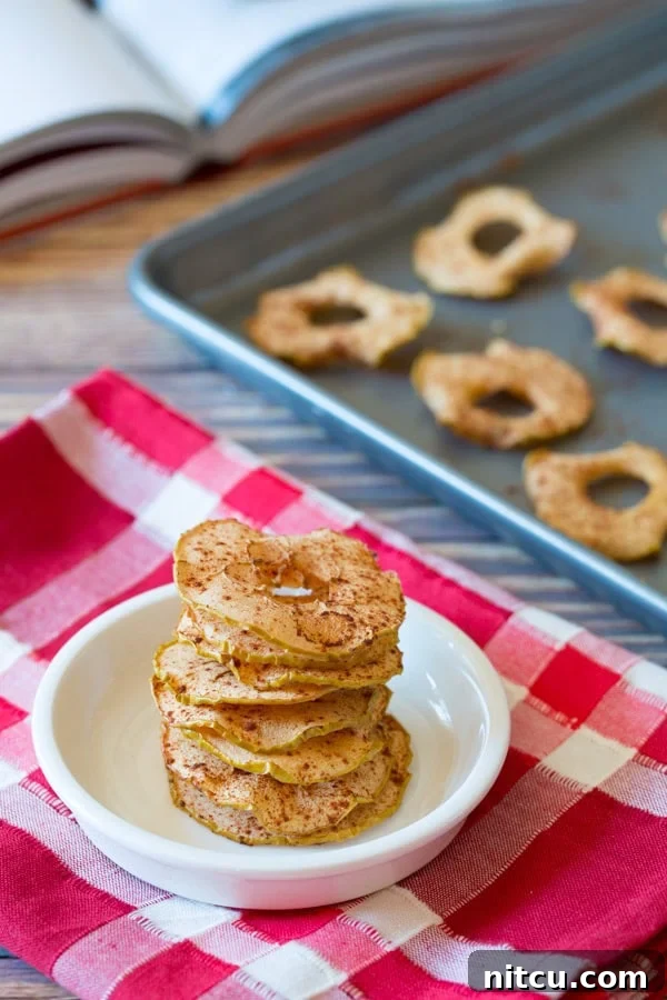 Baked Apple Chips stacked up in a white bowl sitting on a red and white linen. More chips are in the background on a baking sheet.