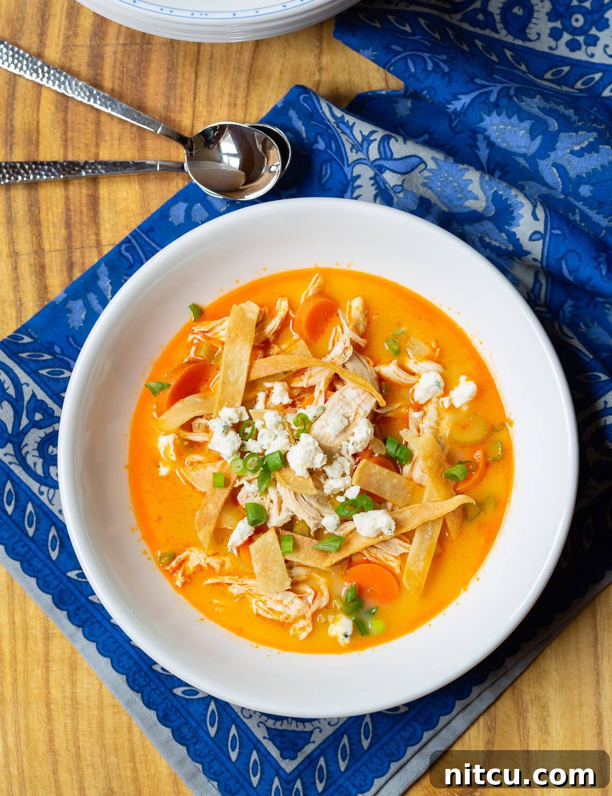 A steaming bowl of Slow Cooker Buffalo Chicken Soup garnished with fresh green onions and crispy tortilla strips, resting on a white surface with a blue cloth.