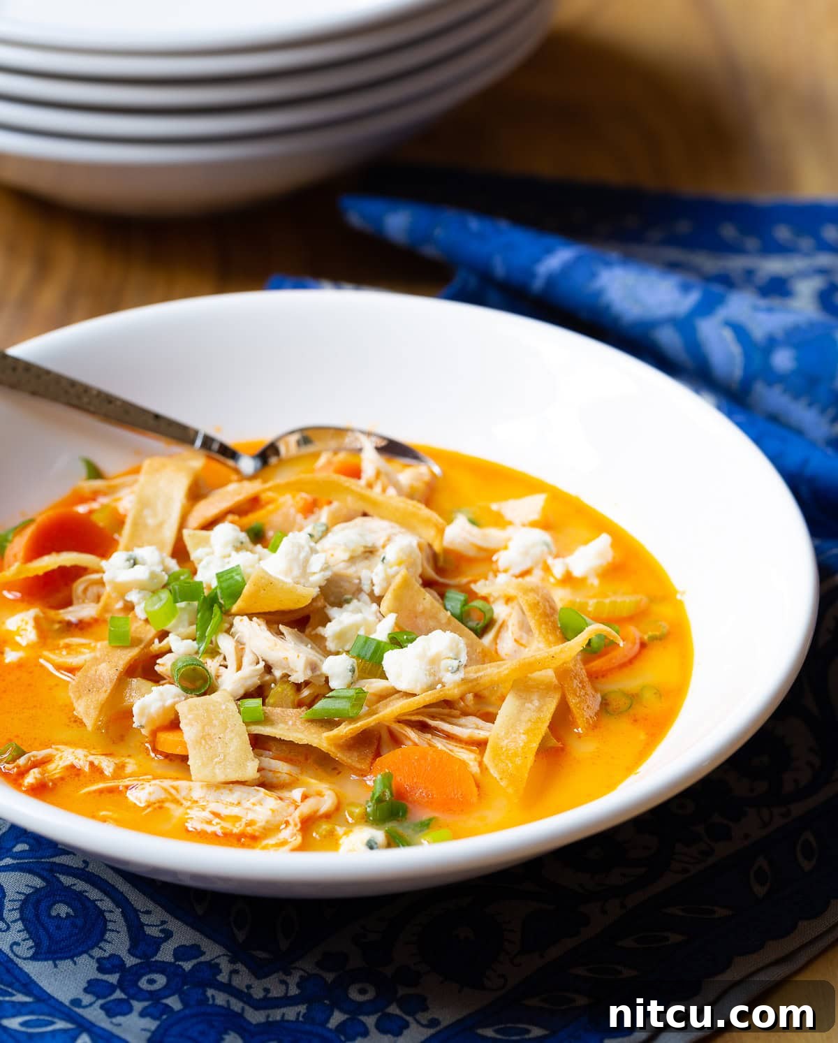 Close-up of a rustic white bowl filled with Easy Buffalo Chicken Soup, topped generously with golden crispy tortilla strips and finely chopped green onions, ready to be enjoyed.