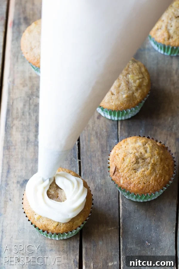 A hand using a piping bag to swirl creamy sour cream frosting onto a baked Hummingbird cupcake.