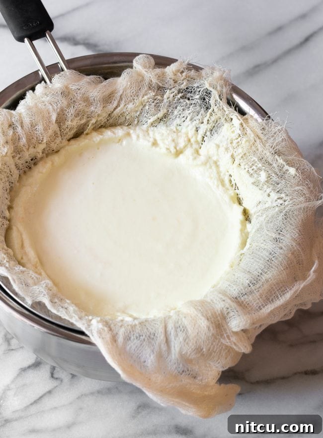 A mound of solidified homemade ricotta cheese resting within the cheesecloth in a sieve, with the whey having drained into the bowl below, showcasing the finished product.