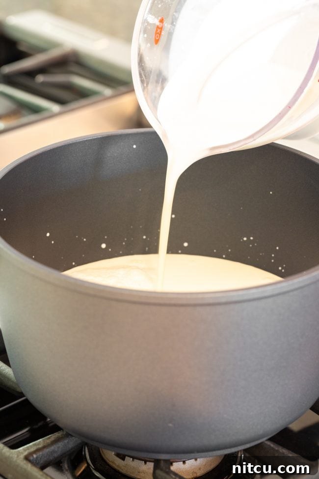 Pouring fresh whole milk from a glass measuring cup into a large metal stock pot, ready for heating.