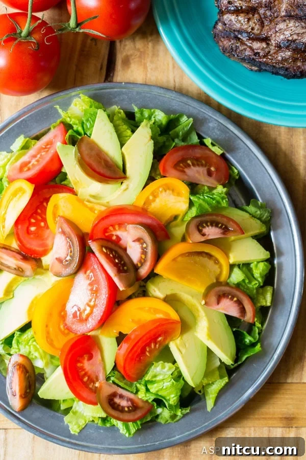 A elegantly plated steak salad, featuring fresh lettuce, vibrant tomatoes, and creamy avocado slices. In the background, thinly sliced grilled steak and additional tomatoes are arranged, hinting at the generous portions and delightful components of the dish.
