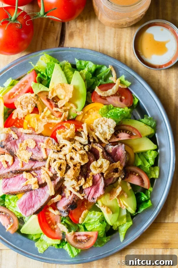 A beautifully composed steak salad on a blue plate, featuring tender steak slices, creamy avocado, ripe tomatoes, and crispy fried onions, all drizzled with a rich vinaigrette. This close-up view emphasizes the freshness and texture of each ingredient.