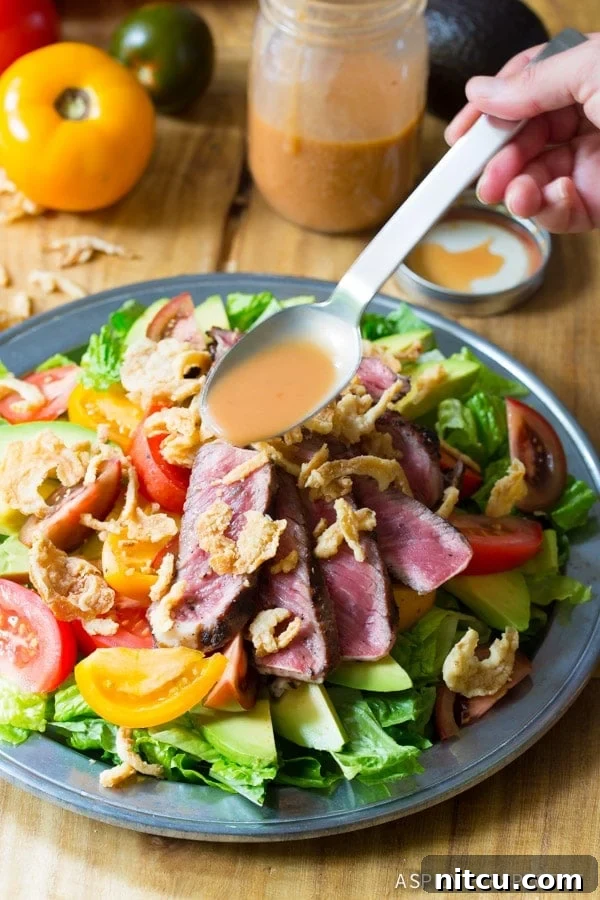 A hand holds a spoon, gracefully drizzling the zesty A1 vinaigrette over a vibrant steak salad. The salad is a colorful medley of tender steak, creamy avocado, juicy tomatoes, and crunchy fried onions, ready to be enjoyed.