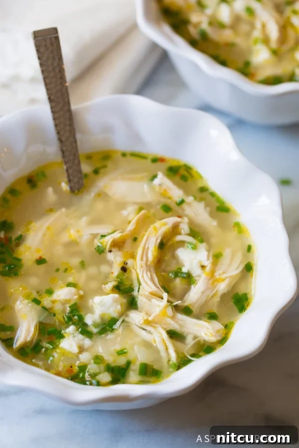 A single white bowl of Greek lemon chicken soup, garnished with chives and a lemon slice, with a spoon nestled inside, ready to be eaten.
