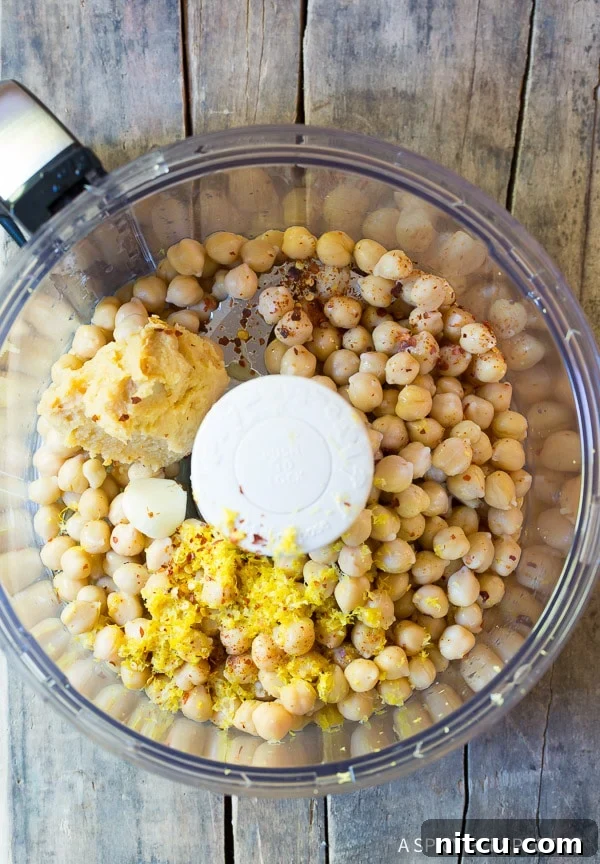 Top down view food processor with chickpeas, lemon zest, scoop of miso paste, and cloves of garlic.
