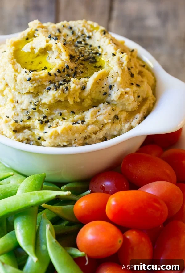 Close view of a white bowl overflowing with homemade miso paste hummus garnished with sesame seeds. Around the dish are fresh sweet peas and grape tomatoes.