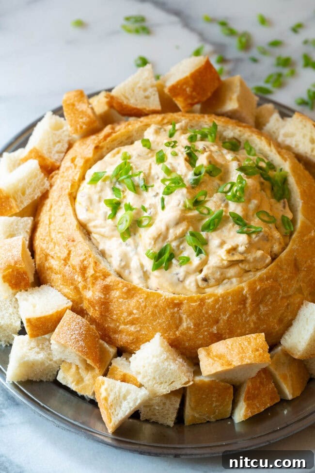 A beautiful sourdough bread bowl brimming with rich, creamy sausage cheese dip, garnished with fresh green onions and surrounded by golden bread cubes for dipping on a rustic wooden platter.