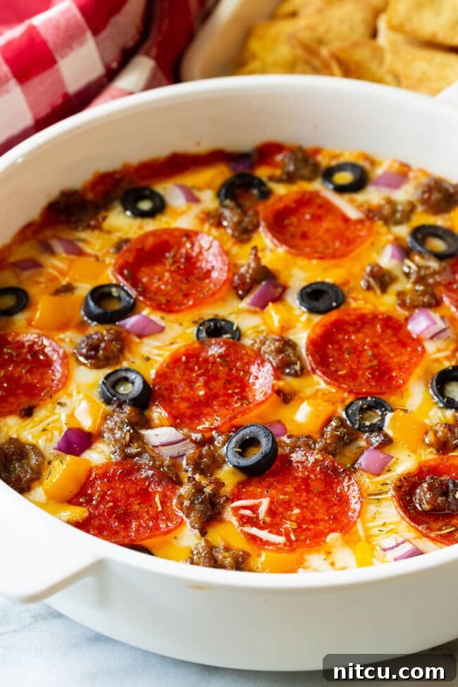 White round dish with loaded supreme pizza dip, topped with pepperoni slices, ground cooked sausage, black olive slices, and chopped red onion.