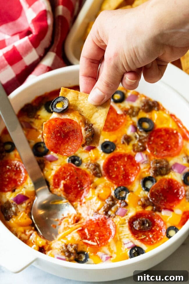 White round dish with loaded supreme pizza dip, topped with pepperoni slices, ground cooked sausage, black olive slices, and chopped red onion. A hand is scooping a bite with a square pita cracker.