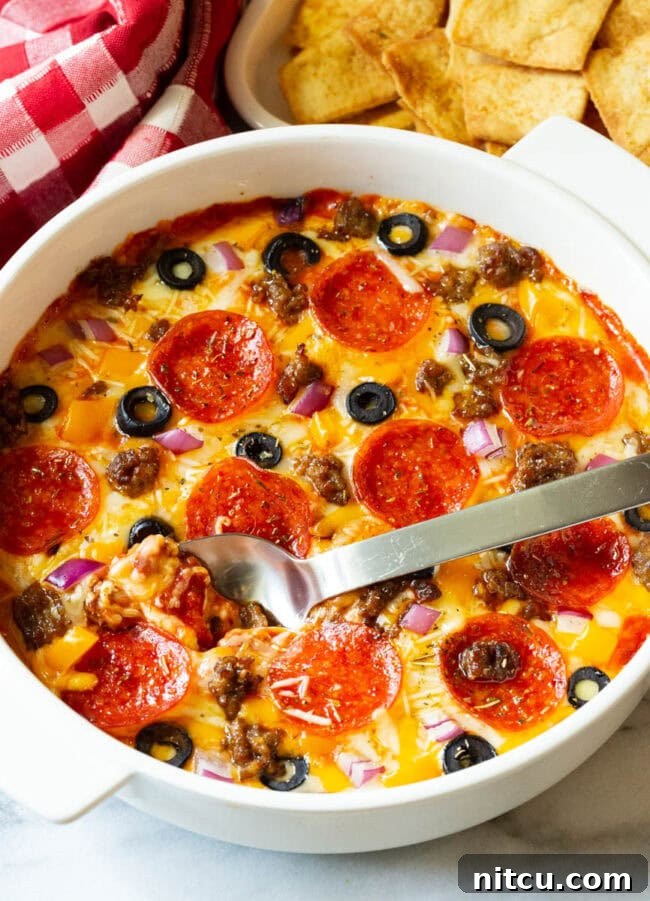 White round dish with loaded supreme pizza dip, topped with pepperoni slices, ground cooked sausage, black olive slices, and chopped red onion.