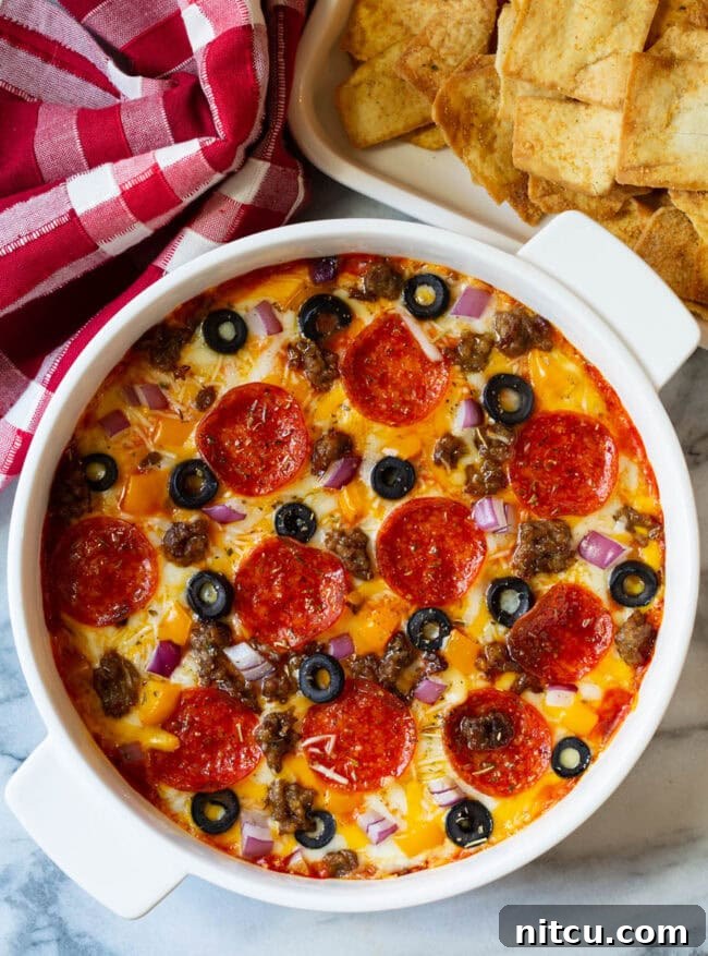 Top down view of a white round dish with loaded supreme pizza dip, topped with pepperoni slices, ground cooked sausage, black olive slices, and chopped red onion.