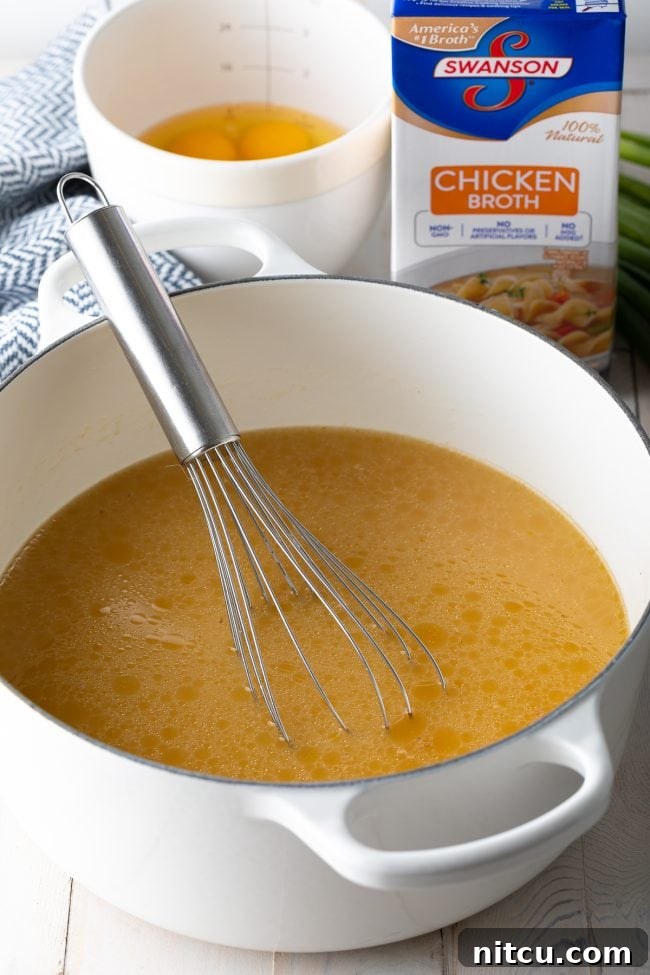 Pot of broth in a large white cooking pot, with a metal whisk in the pot. In the background are a box of chicken broth and a white mixing bowl with eggs.