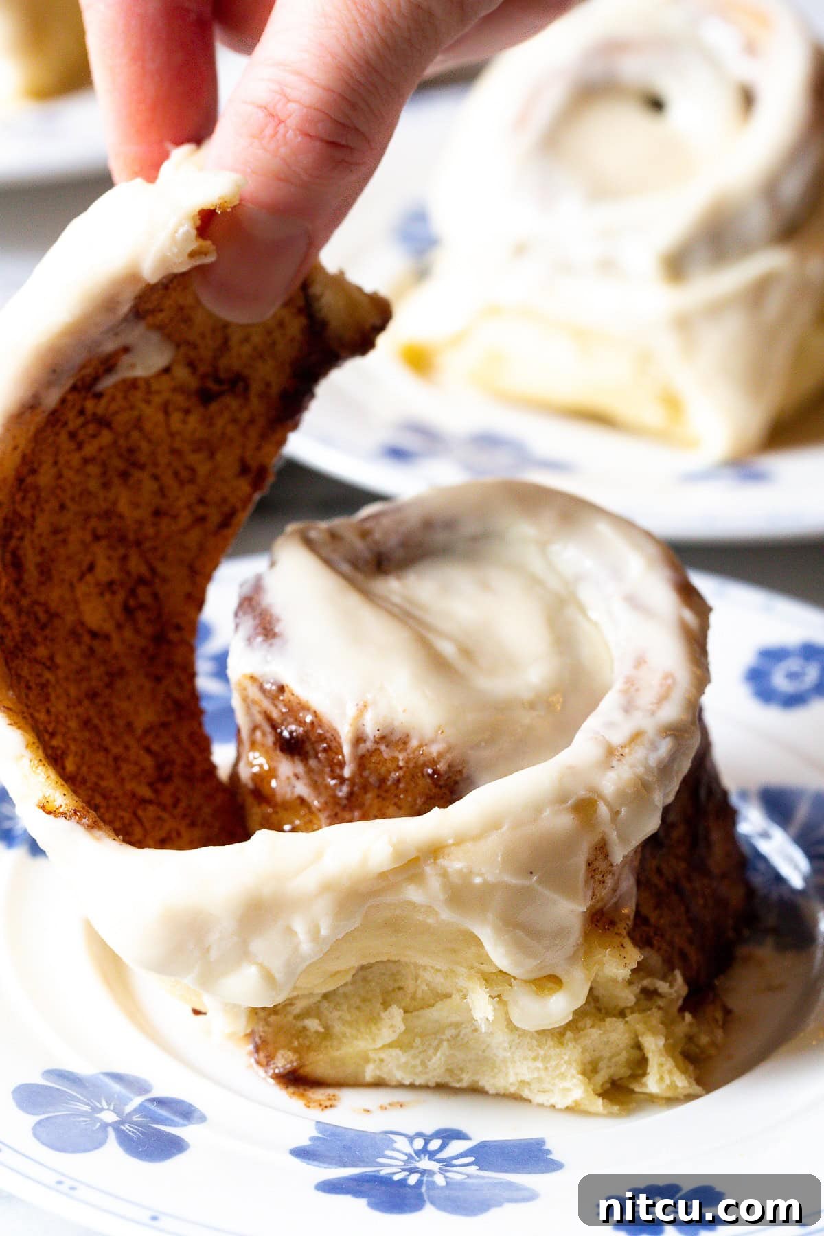 A hand gently pulling apart a warm, freshly baked cinnamon roll on a plate, revealing its soft, spiraled interior.