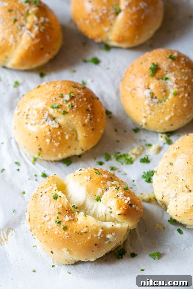 One garlic knot ripped to show its fluffy interior, with the rest of the batch lying behind it on a baking sheet.