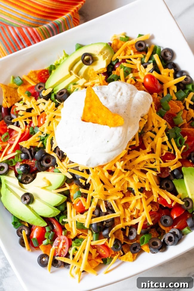A vibrant Dorito taco salad, richly loaded with crisp lettuce, halved grape tomatoes, thinly sliced black olives, chopped green onions, creamy avocado slices, and generous shredded cheddar cheese, all topped with a dollop of cool ranch sour cream and a signature Dorito chip.