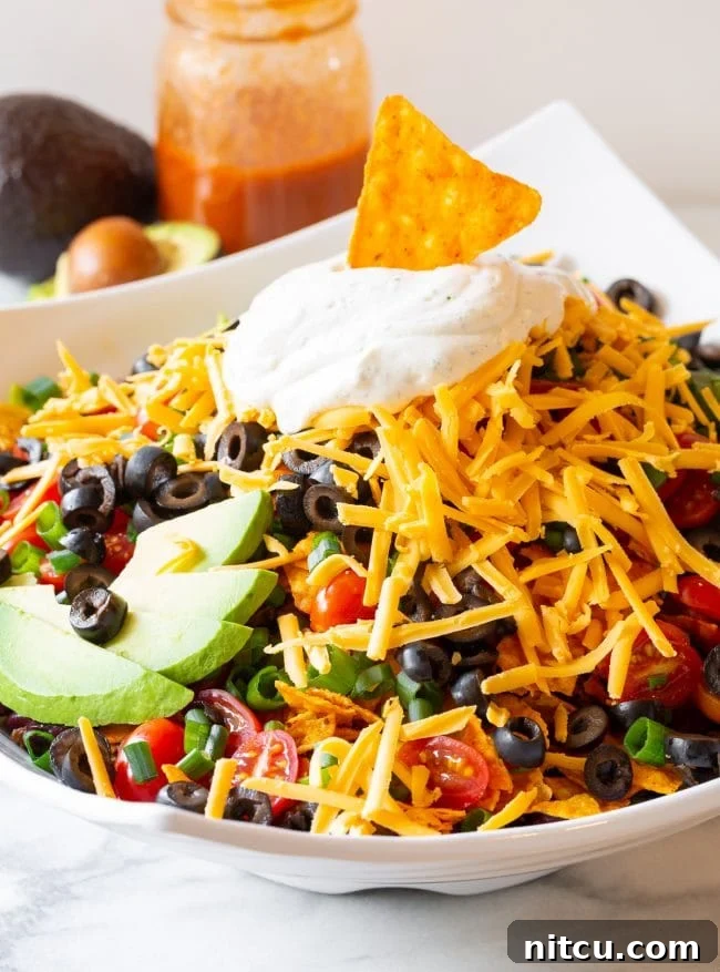 A close-up of a generously loaded Dorito taco salad bowl, showcasing crisp lettuce, vibrant grape tomatoes, sliced black olives, green onions, avocado, shredded cheddar, and dollops of ranch sour cream, with a Dorito chip serving as a garnish.