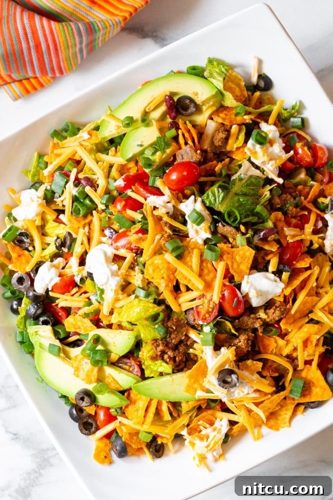 A beautifully arranged Dorito taco salad, overflowing with crisp lettuce, vibrant grape tomatoes, black olives, green onions, avocado slices, and shredded cheddar cheese, adorned with multiple dollops of creamy ranch sour cream around the edges.