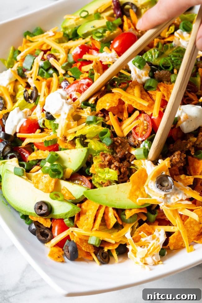 A hand using wooden tongs to gently toss a large Dorito taco salad, revealing layers of crushed Dorito chips and fresh salad fixings.