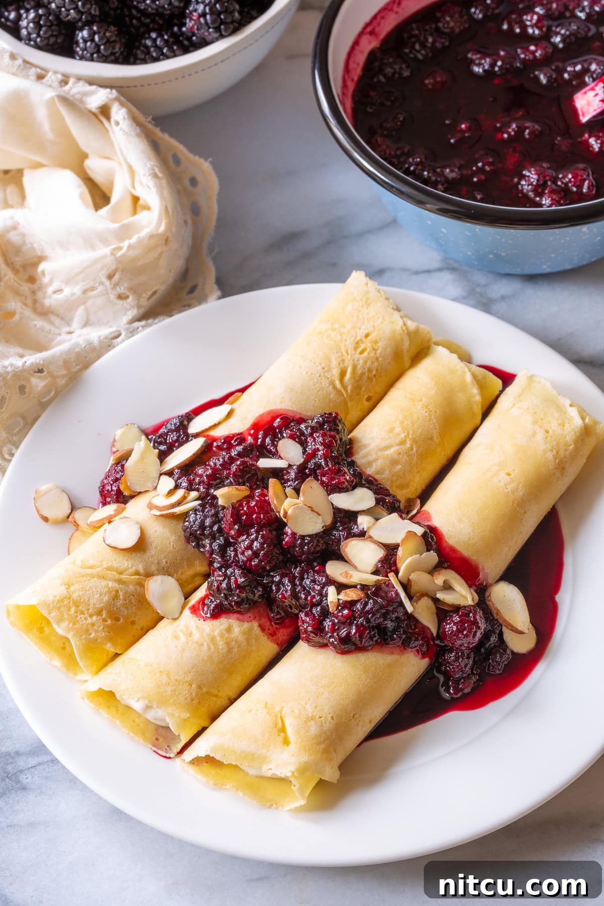 Blintz pancakes resting on a white plate with blackberry sauce, garnished with almonds
