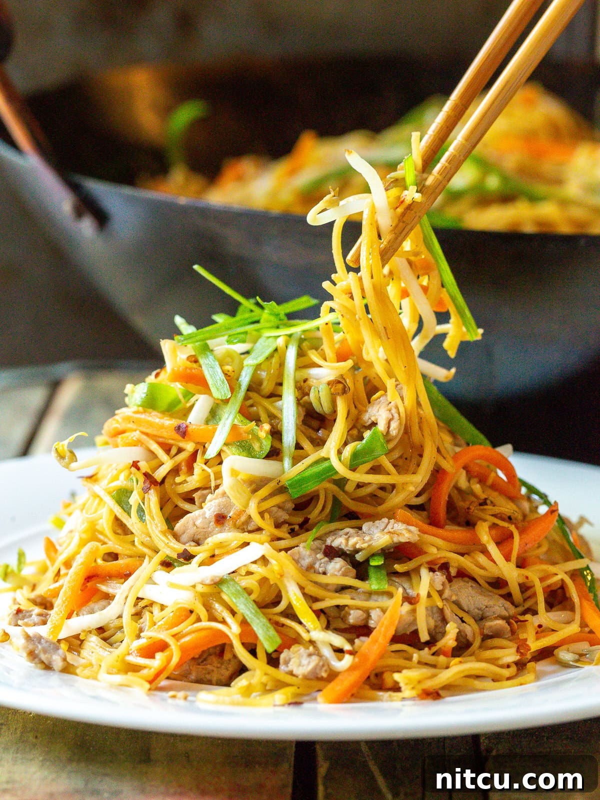 A close-up shot of Cantonese Lo Mein generously piled on a white plate, with a pair of chopsticks picking up a mouthful of noodles, pork, and vegetables.