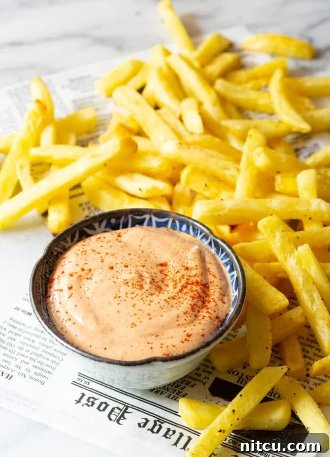 A vibrant top-down view of Southern Comeback Sauce in a bowl, flanked by golden French fries and laid out on a sheet of newspaper for a rustic feel.