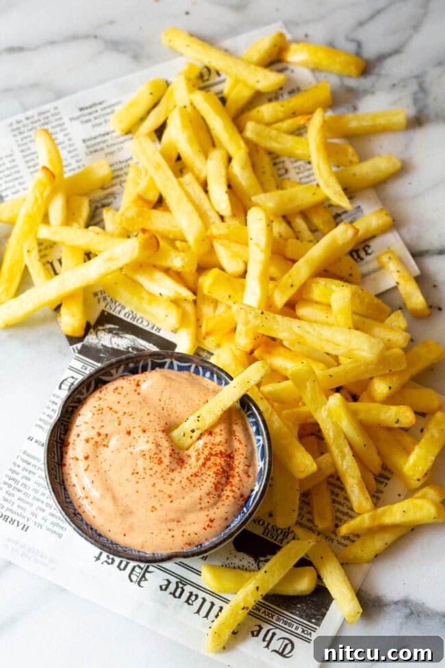 A hand dipping a crispy French fry into a bowl of luscious Southern Comeback Sauce, with more fries and a newspaper background.