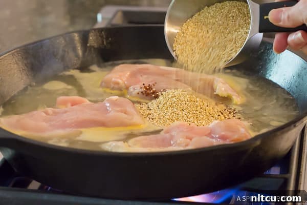 Adding uncooked quinoa to the pan with chicken, garlic, and chicken broth, preparing for it to simmer and absorb the rich flavors in the Healthy One-Pot Chicken Broccoli Quinoa.
