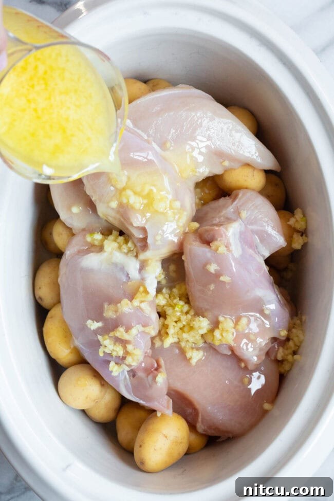 Raw chicken and potatoes arranged in a slow cooker, with a luscious garlic butter mixture being generously poured over the ingredients.