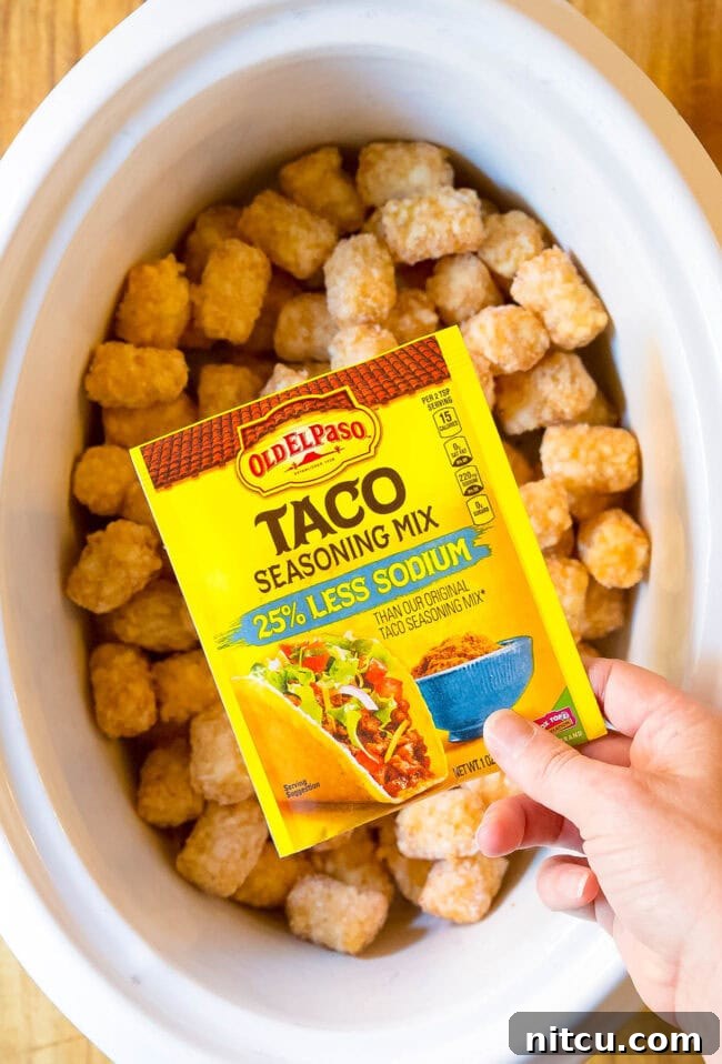 Frozen tater tots in a baking dish with a hand holding a packet of taco seasoning above it. 
