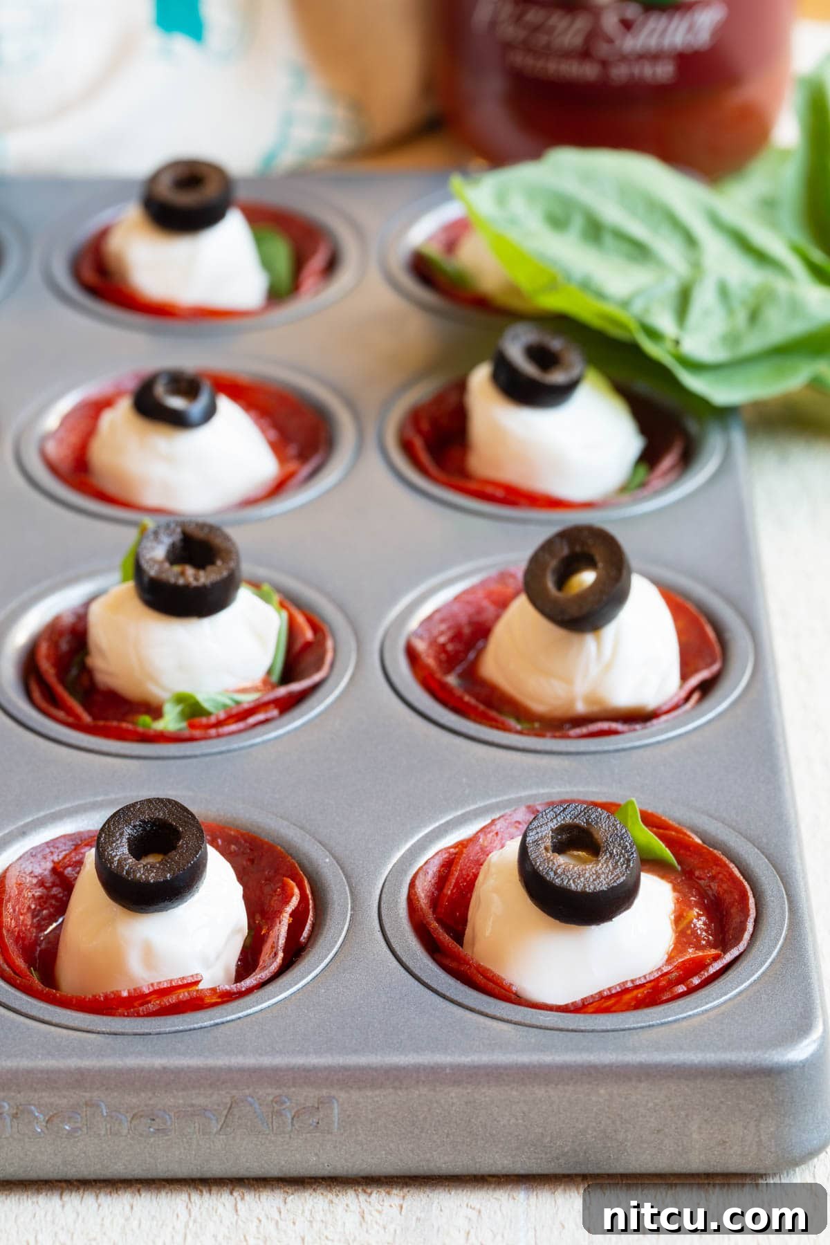 A mini muffin pan showcases pepperoni cups, each meticulously filled with fresh basil, zesty pizza sauce, a mozzarella ball, and sliced olives, eagerly awaiting their final bake.