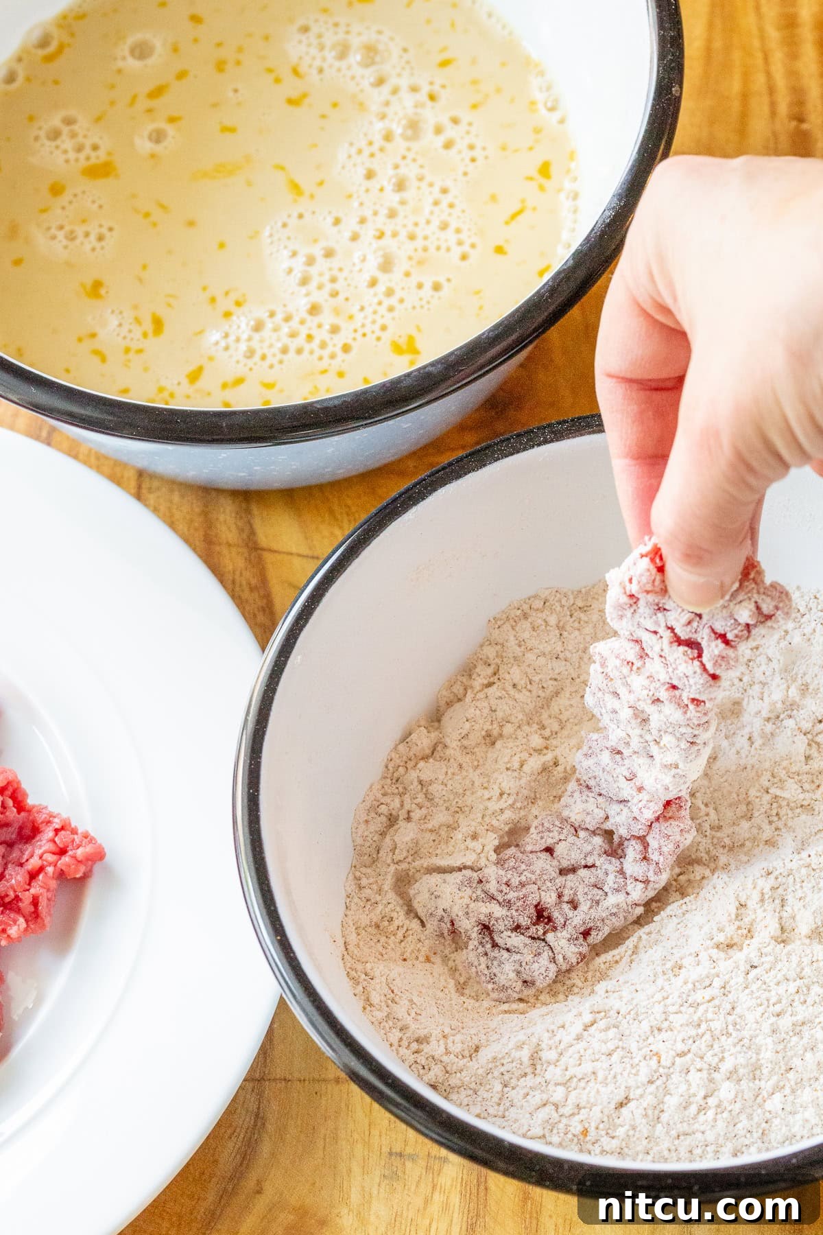 A hand dipping a strip of raw beef into a bowl of batter and then into a bowl of seasoned flour, preparing it for frying.