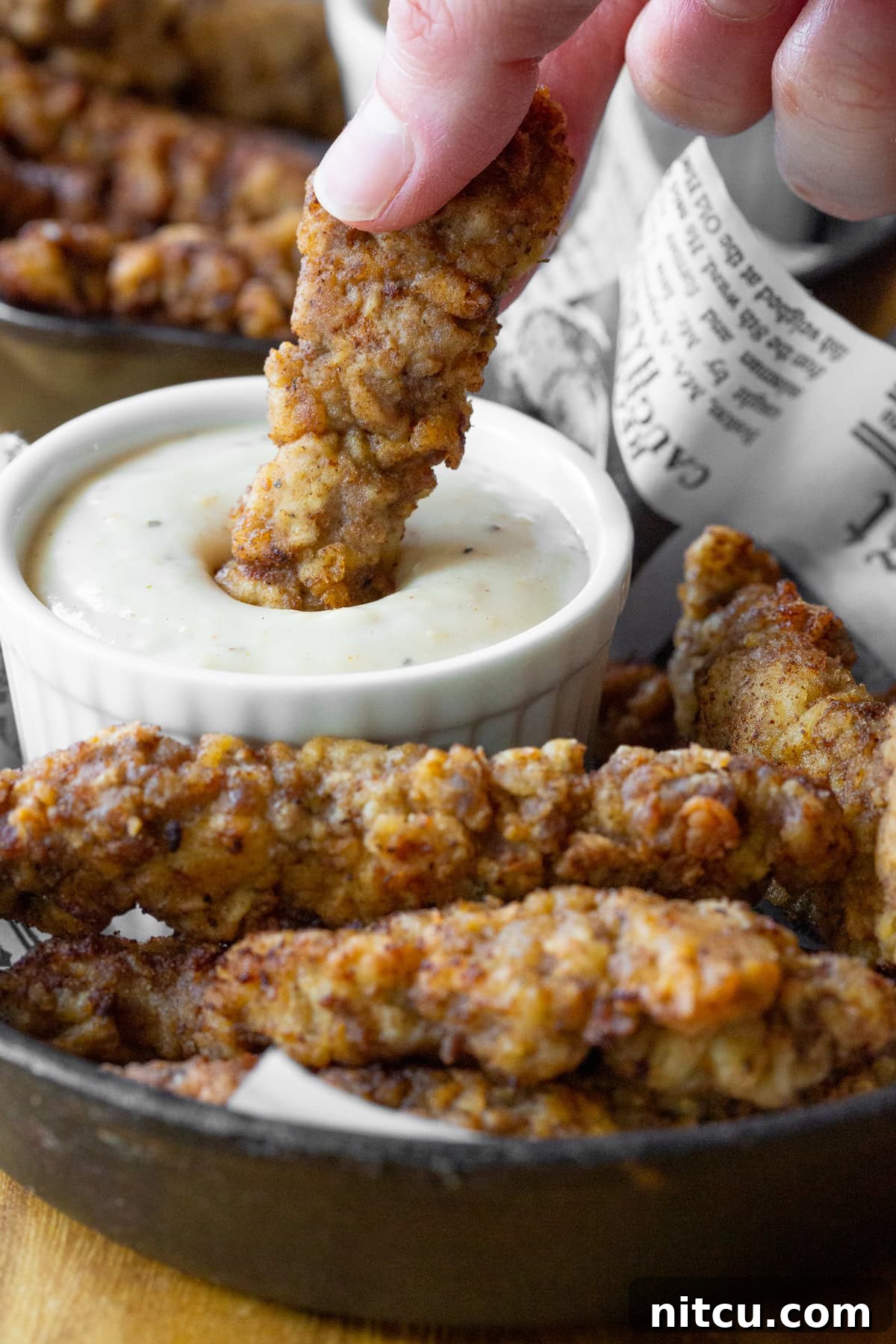 A hand dipping a crispy steak finger into a small ramekin of creamy country gravy, highlighting the perfect dipping experience.