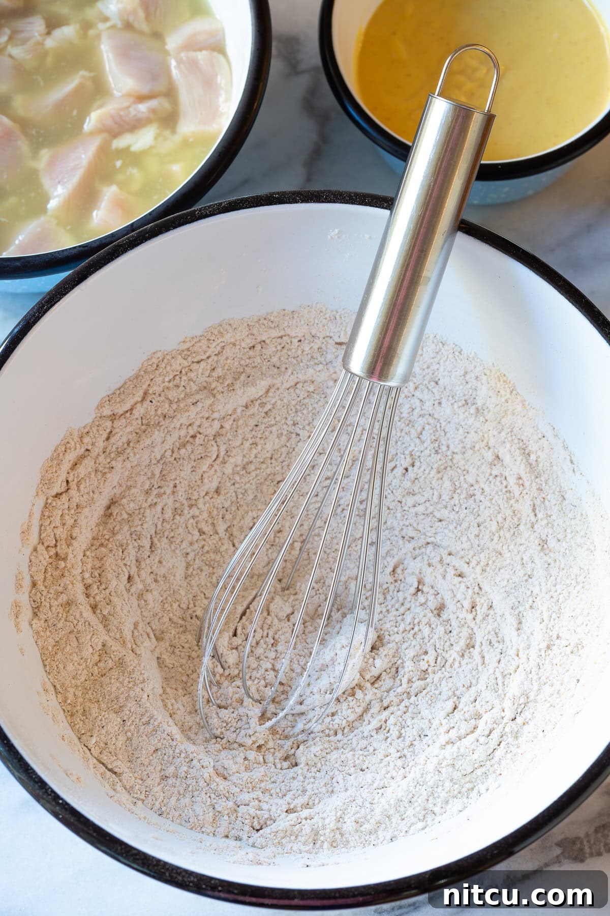 A whisk in a bowl of flour mixture, with other spice bowls in the background, preparing the dry dredge.