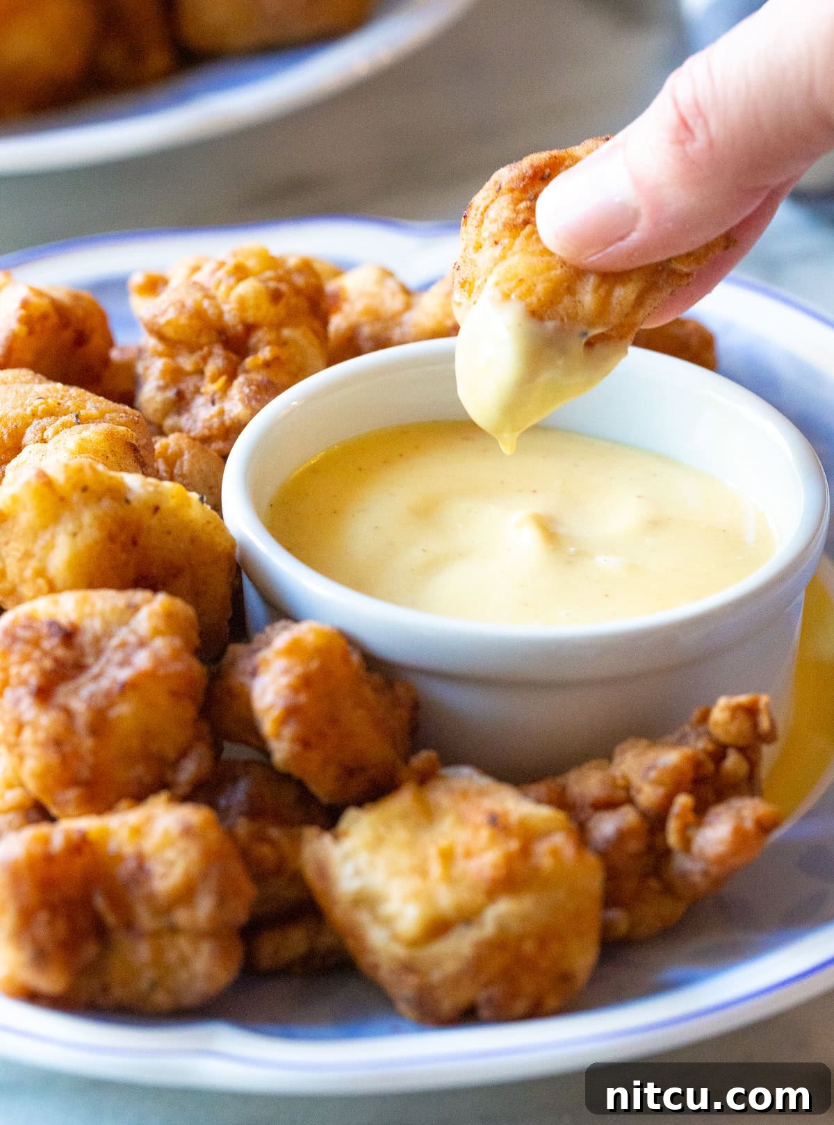 A hand dipping a crispy chicken nugget into a small bowl of sauce, surrounded by more golden nuggets.