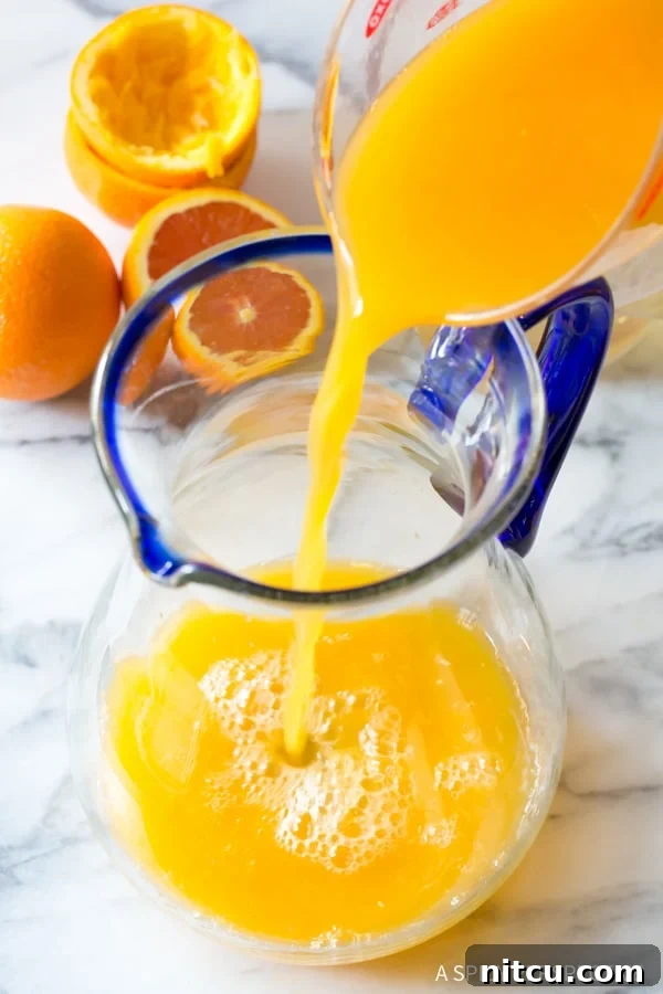 Freshly squeezed orange juice, a rich golden hue, being carefully poured into a large clear glass pitcher, symbolizing the start of a homemade orangeade batch.