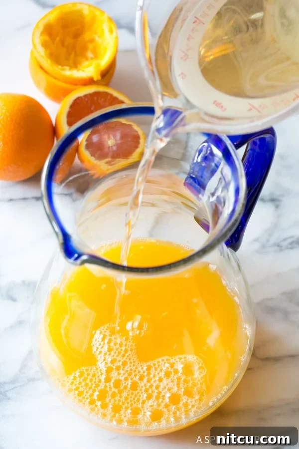 A stream of clear simple syrup being poured into a large glass pitcher containing citrus juices, an essential step in preparing homemade orangeade.