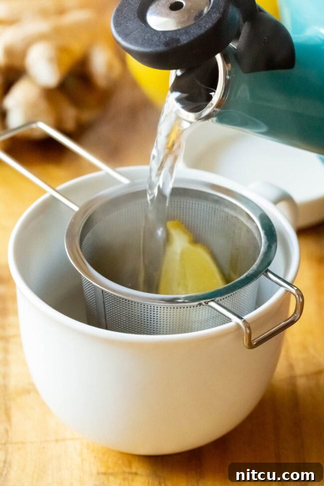 A clear mug showing hot water being poured over lemon and grated ginger held in a tea infuser, creating a vibrant yellow hue.