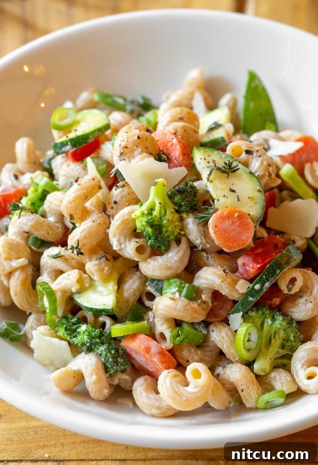A beautifully presented white bowl of skinny pasta primavera, garnished with fresh zucchini, carrots, broccoli, red peppers, and a sprinkle of shaved parmesan cheese.