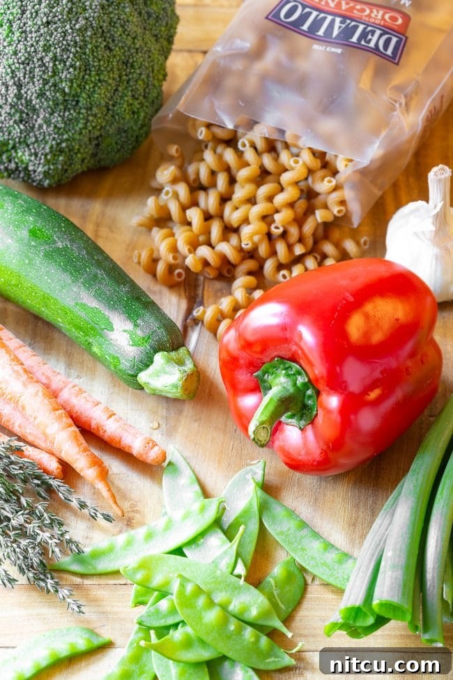 A collection of fresh recipe ingredients laid out: a whole red bell pepper, fresh zucchini, vibrant carrots, crisp snow peas, green onions, raw spiral pasta (fusilli), and a bulb of garlic.