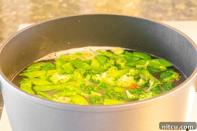 A large pot of boiling water with broccoli florets, snow peas, and sliced carrots, showing the blanching process.